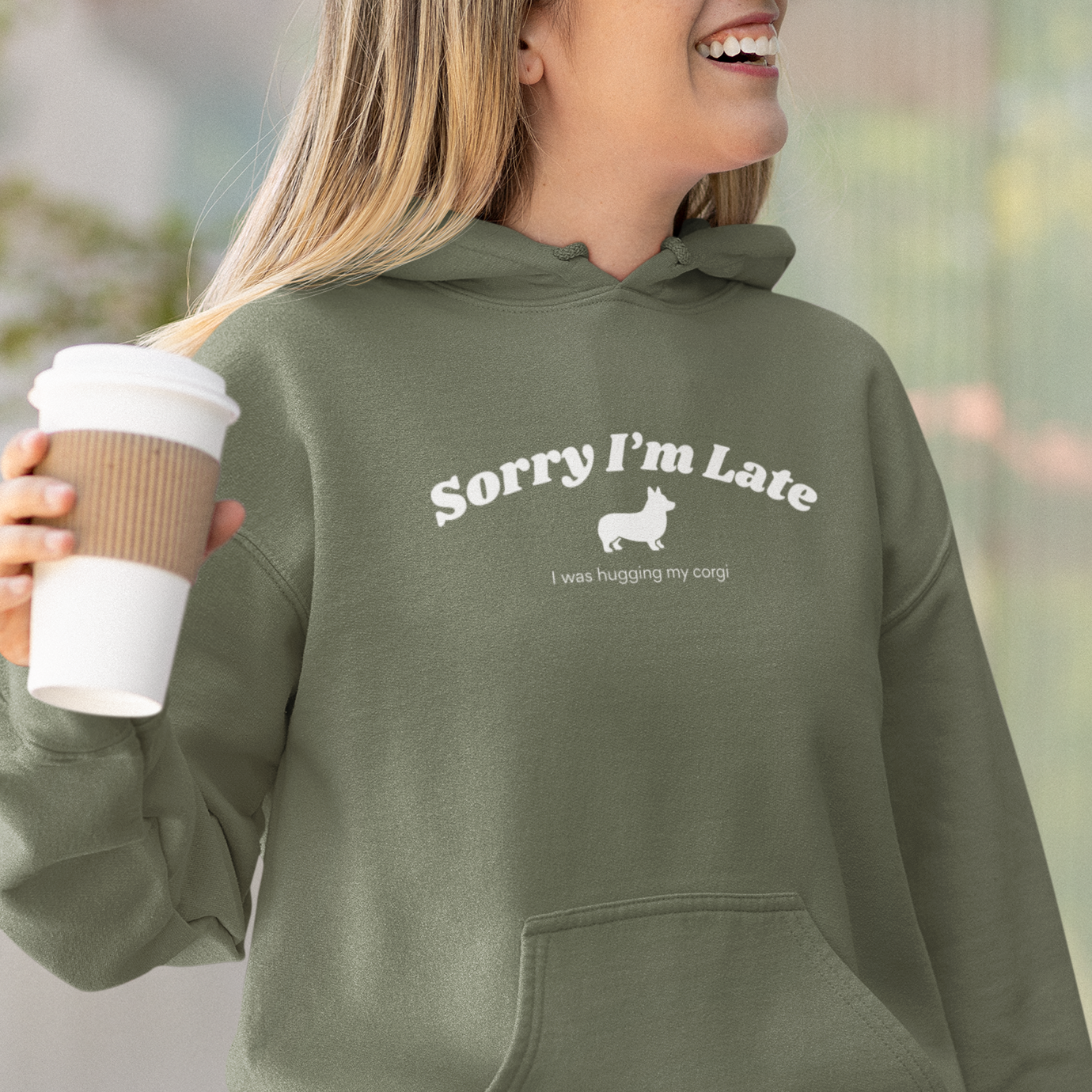 Woman wearing a green hoodie with text and a dog graphic, holding a coffee cup outdoors.
