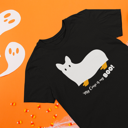 A black short-sleeve T-shirt laid flat on an orange background, featuring a white ghost-shaped dog graphic with yellow paws and the text 'My Cat Is my BOO!'. White ghost cutouts, pumpkins, and candy corn are arranged around the shirt as seasonal props.